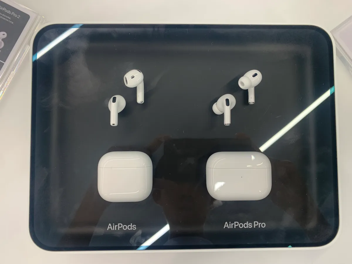 AirPods 4 on the left and AirPods Pro 2 on the right, each with its white charging case, arranged on a dark store display tray