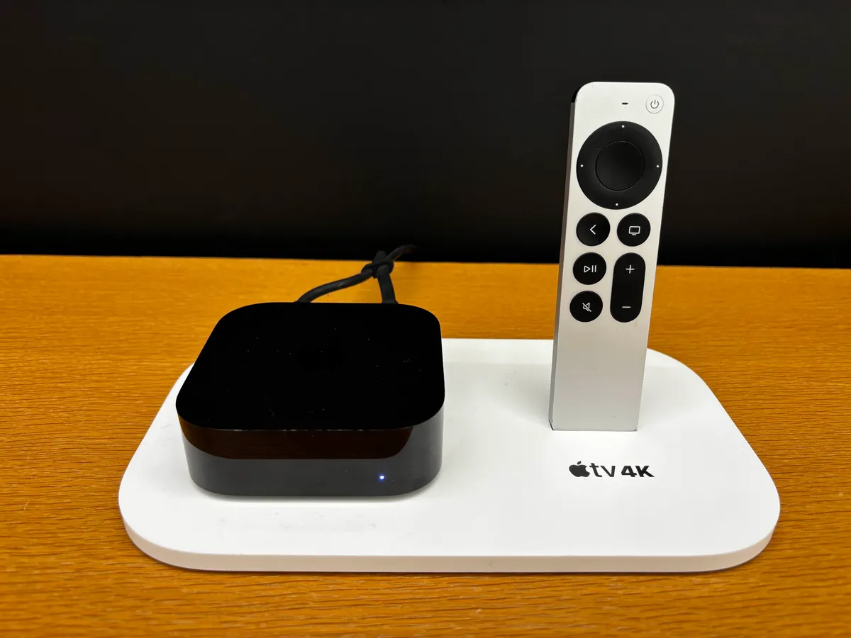 An Apple TV 4K box and a silver Siri Remote on a white stand with the Apple TV 4K logo, in front of a black wall