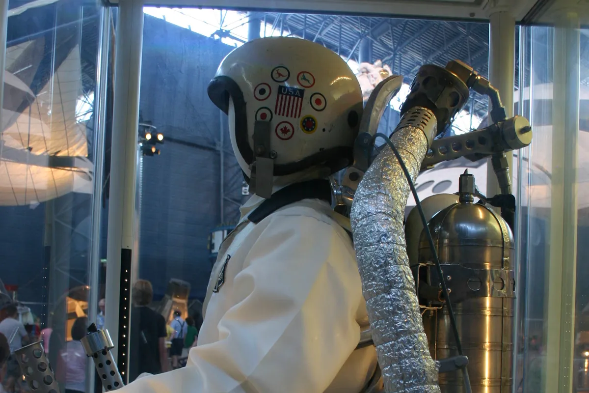 A white Bell Rocket Belt No. 2 flight suit with helmet, silver insulated tube and cylindrical fuel tank on the back, shown behind glass in a museum