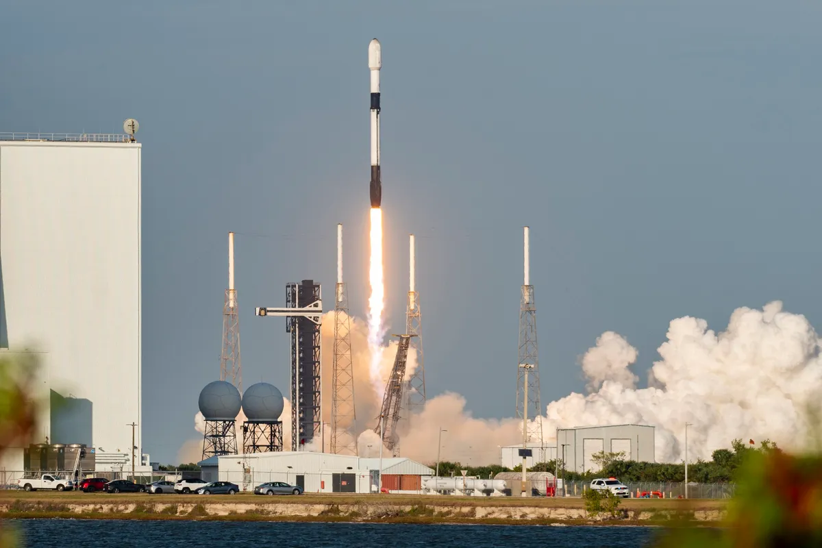 A white and black Falcon 9 rocket lifting off from a launch pad at Cape Canaveral on a column of flame, with a large white cloud of exhaust to the right and a lagoon in the foreground