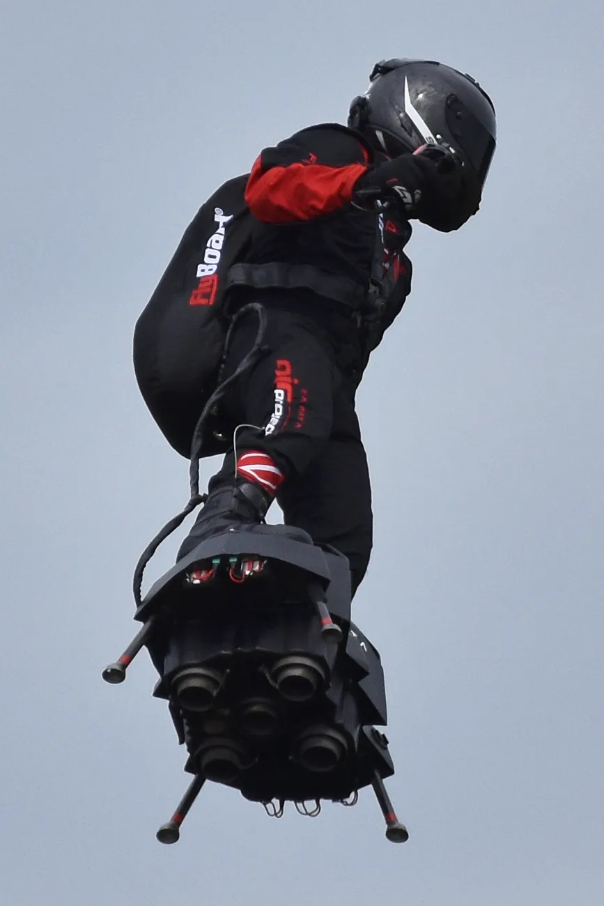 A pilot in a black helmet and black suit flying on a Flyboard Air, seen from below, with the jet engine nozzles under the board and a fuel backpack behind the pilot