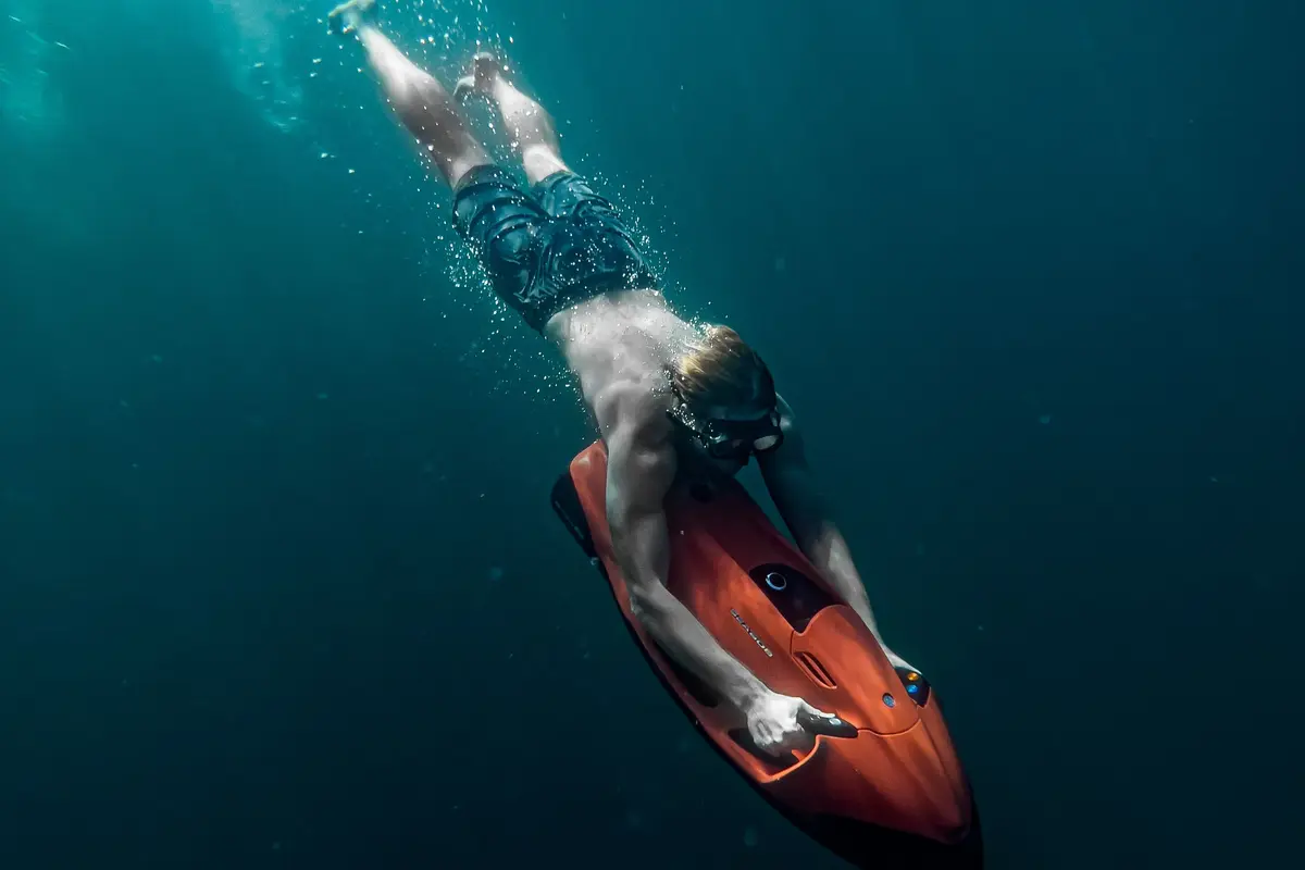 A freediver gliding head-first through dark teal water on an orange board-style sea scooter, holding its handles with both hands while bubbles rise behind