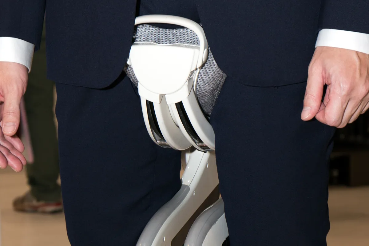 Close-up of a white and gray hip-mounted walking assist frame with two curved legs, worn between the legs of a person in a dark suit