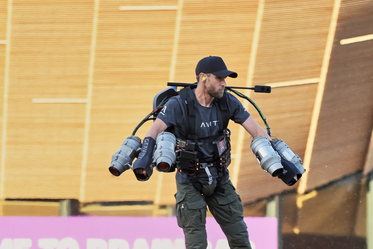 Richard Browning hovering a few feet above the ground in a Gravity jet suit, with two small jet engines on each arm and a backpack, while a crowd films him