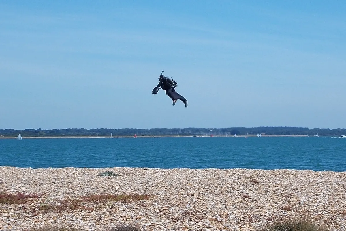 A jet suit pilot flying low over the sea beside a pebble beach, small against a blue sky, with sailboats on the water behind