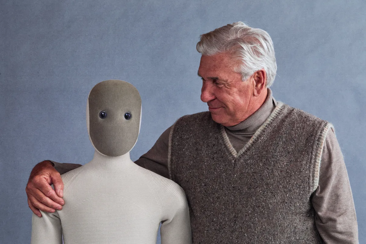 NEO, a humanoid robot with a soft knit body and a smooth gray face with two round eyes, standing next to a smiling older man who rests an arm on its shoulder