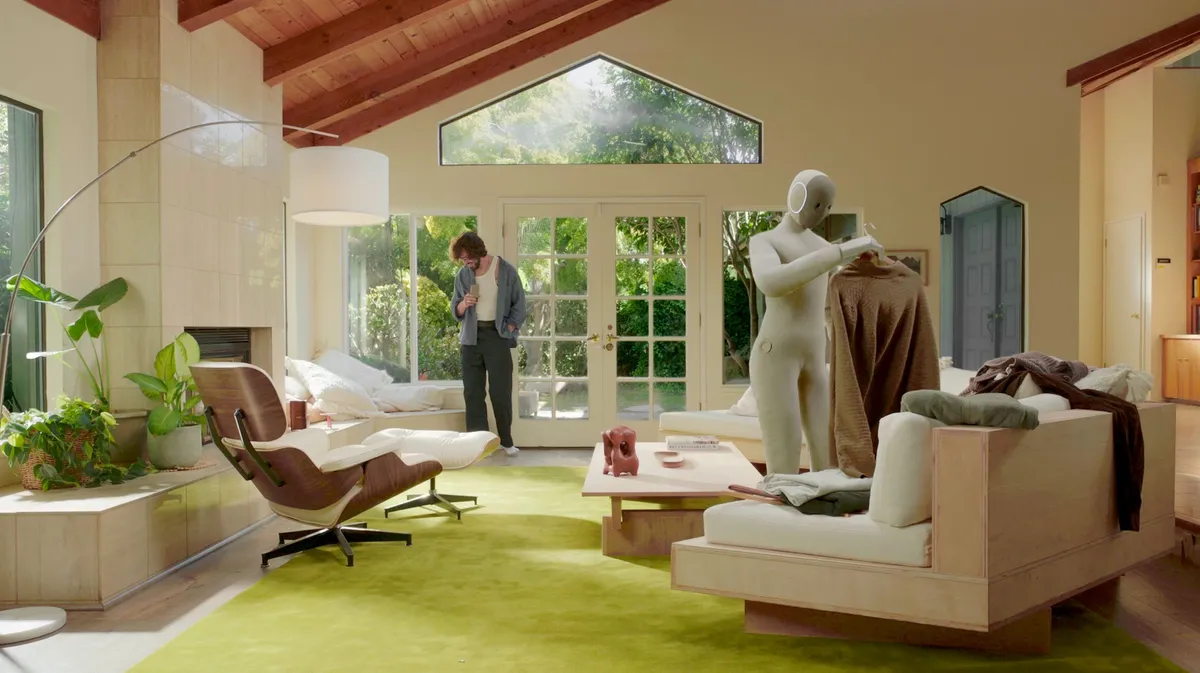 NEO, a light-colored humanoid robot, holding a garment on a hanger in a sunny living room while a man walks in the background