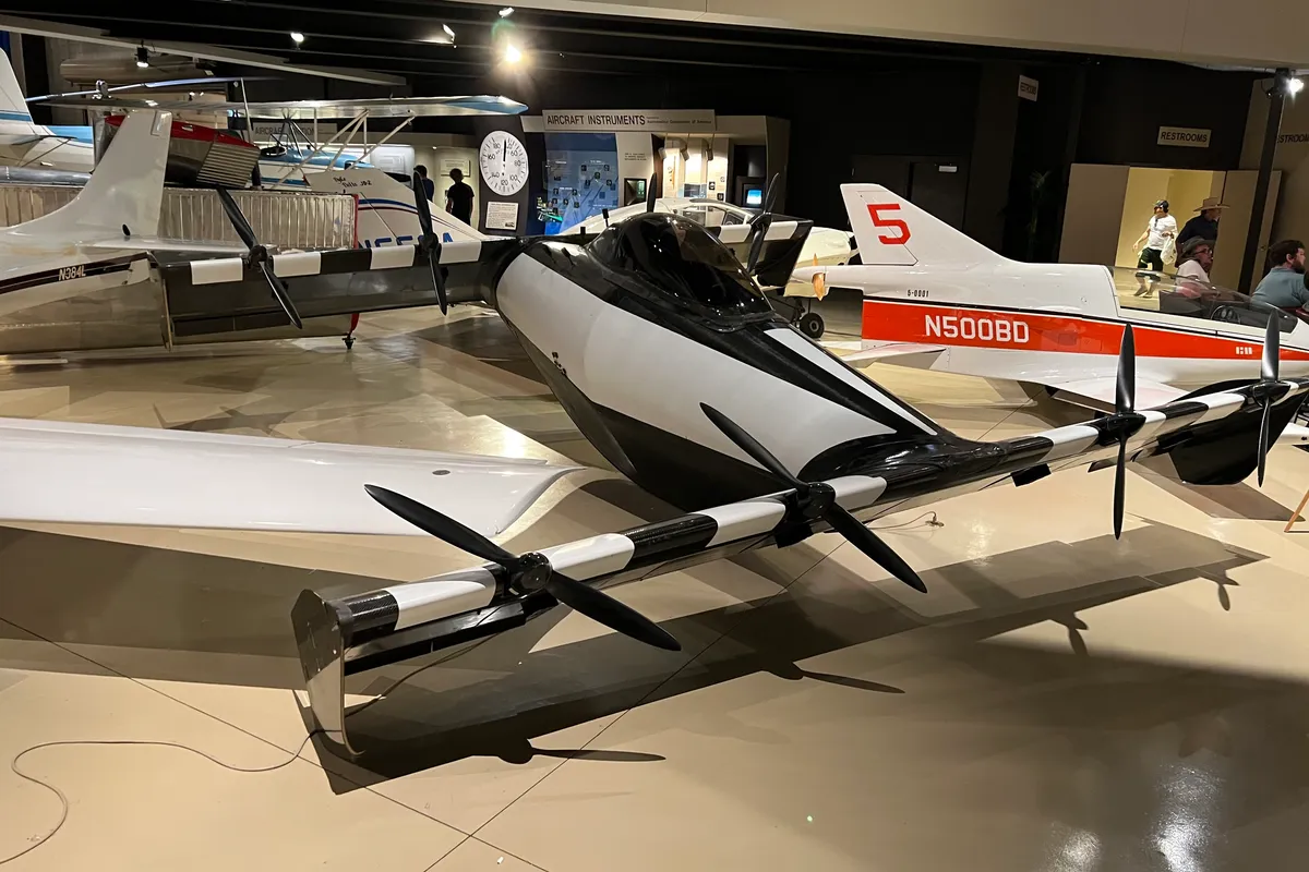 A black and white Opener BlackFly single-seat electric aircraft on a museum floor, with a bubble canopy and propellers along two thin wings