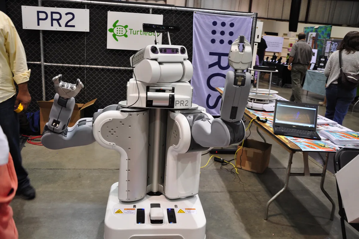 PR2, a white two-armed research robot with grippers raised, standing at a technology fair booth