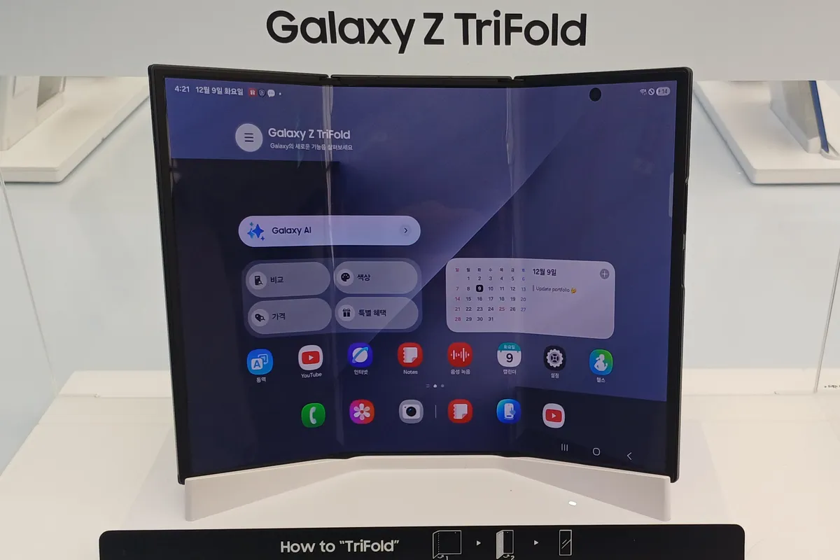 A Samsung Galaxy Z TriFold unfolded on a white display stand in a store, with a home screen showing across its three connected panels under a Galaxy Z TriFold sign