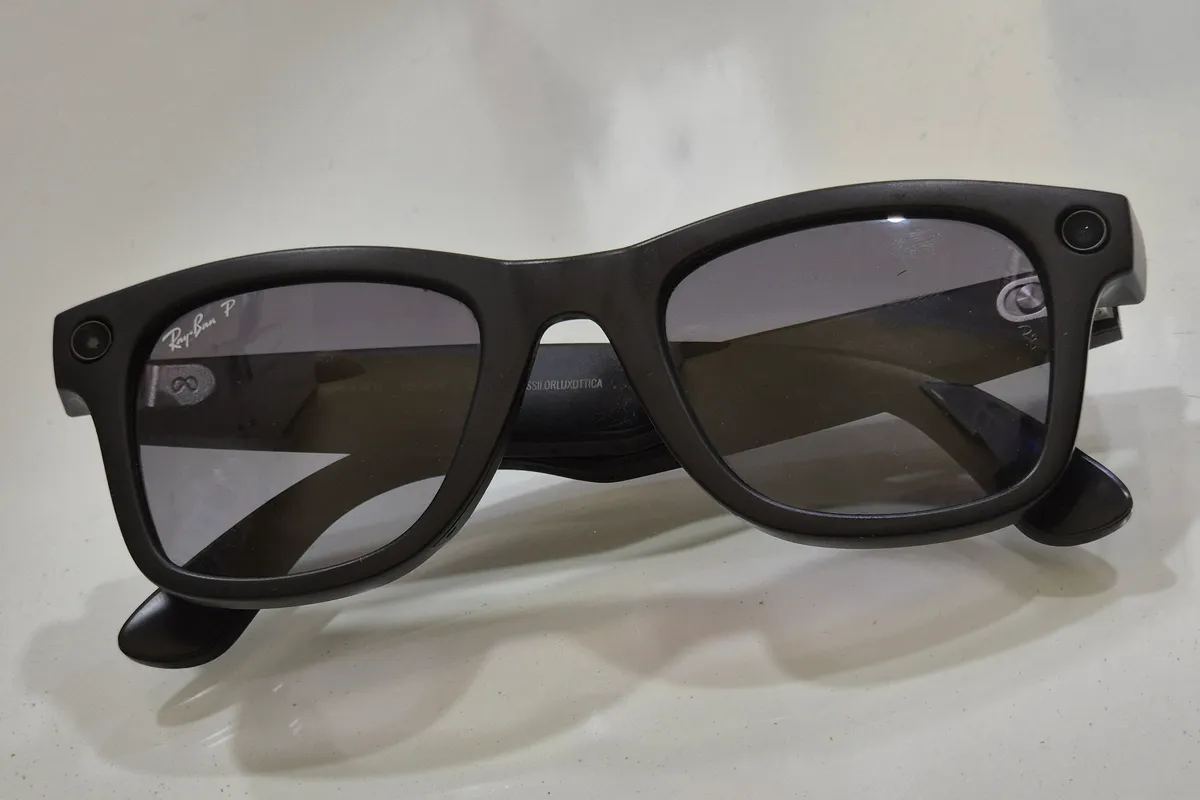 Black Ray-Ban Meta Wayfarer smart glasses with a small camera lens at each outer corner of the frame, lying on a white surface