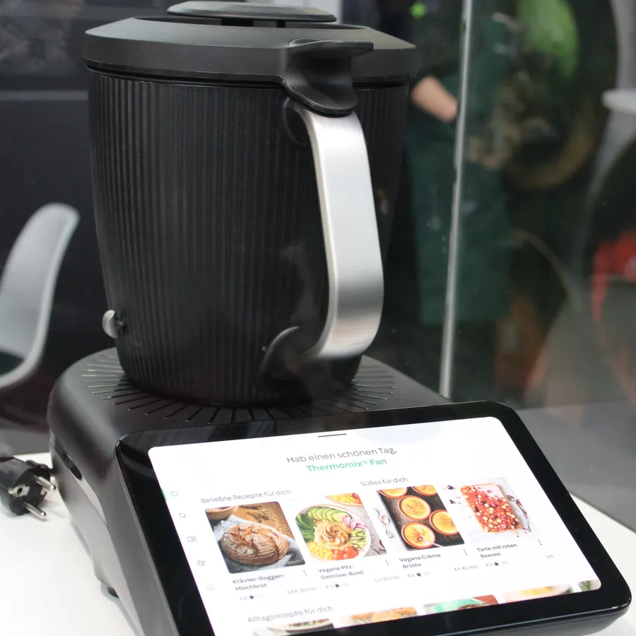 Black Thermomix TM7 cooking bowl with a silver handle on a base with a tilted touchscreen showing recipe photos