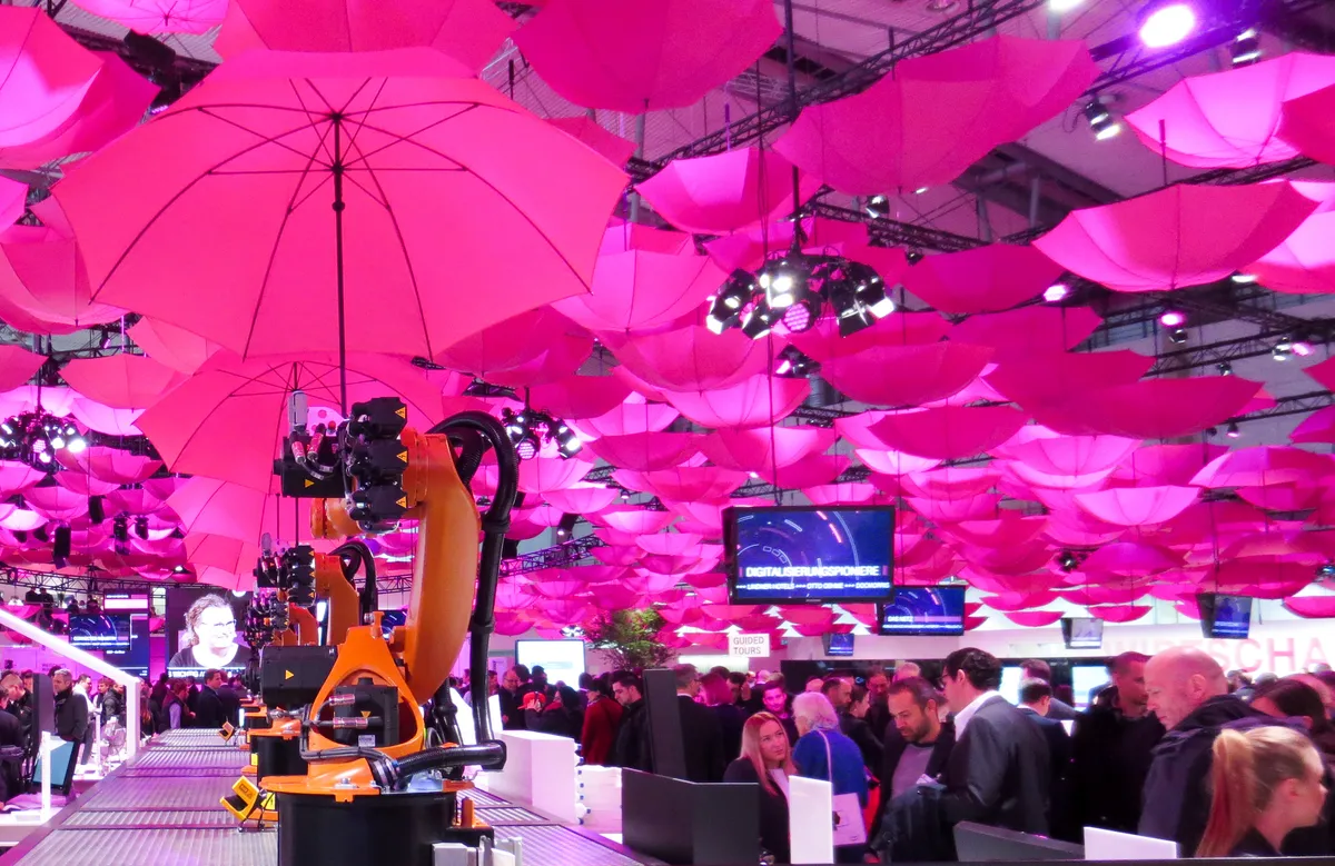 An orange industrial robot arm at a trade fair booth under a ceiling of glowing pink umbrellas, with visitors in the foreground