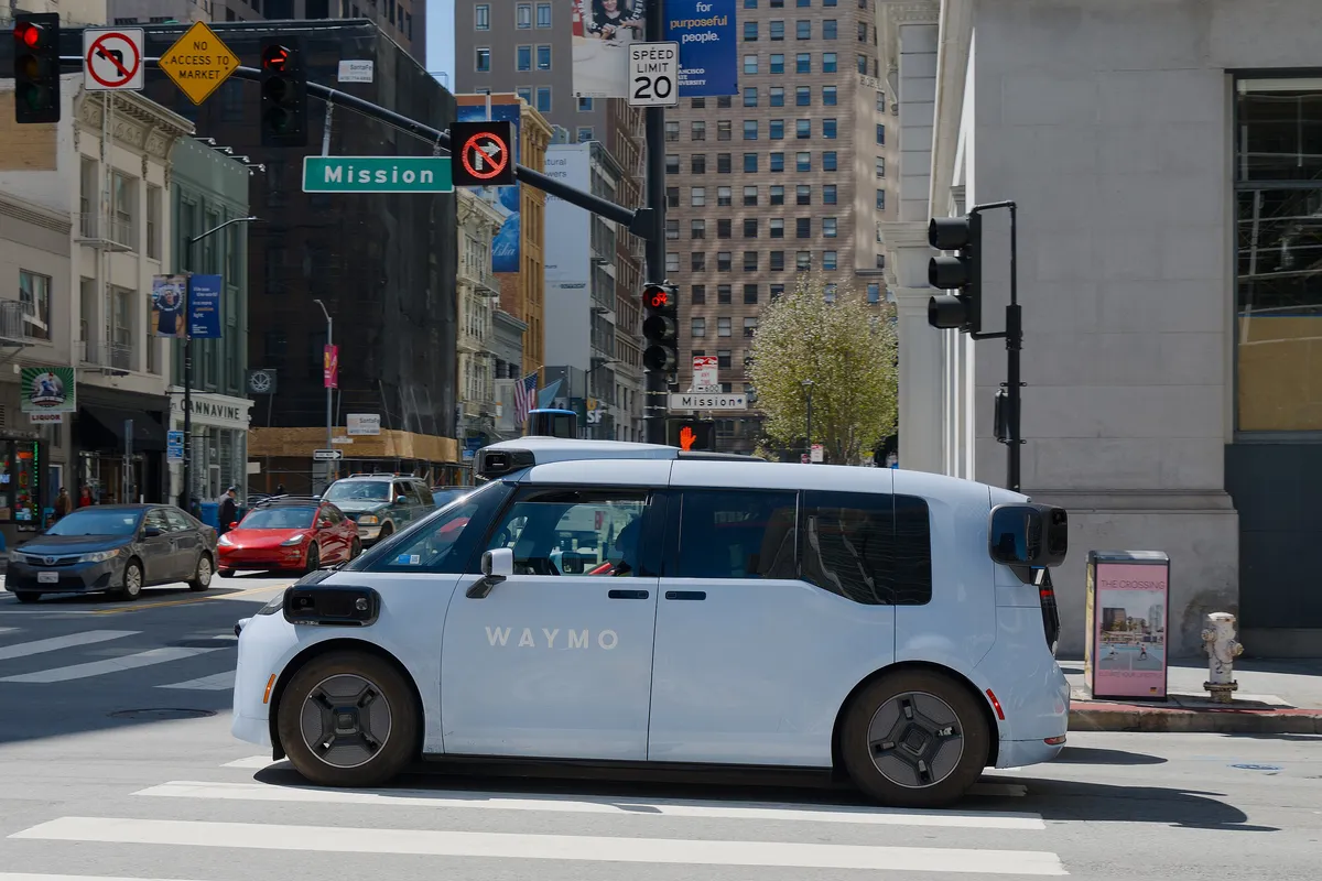 A light blue Waymo Ojai robotaxi, a boxy electric van with the word Waymo on the door, stopped at a crosswalk at the corner of Mission Street in downtown San Francisco