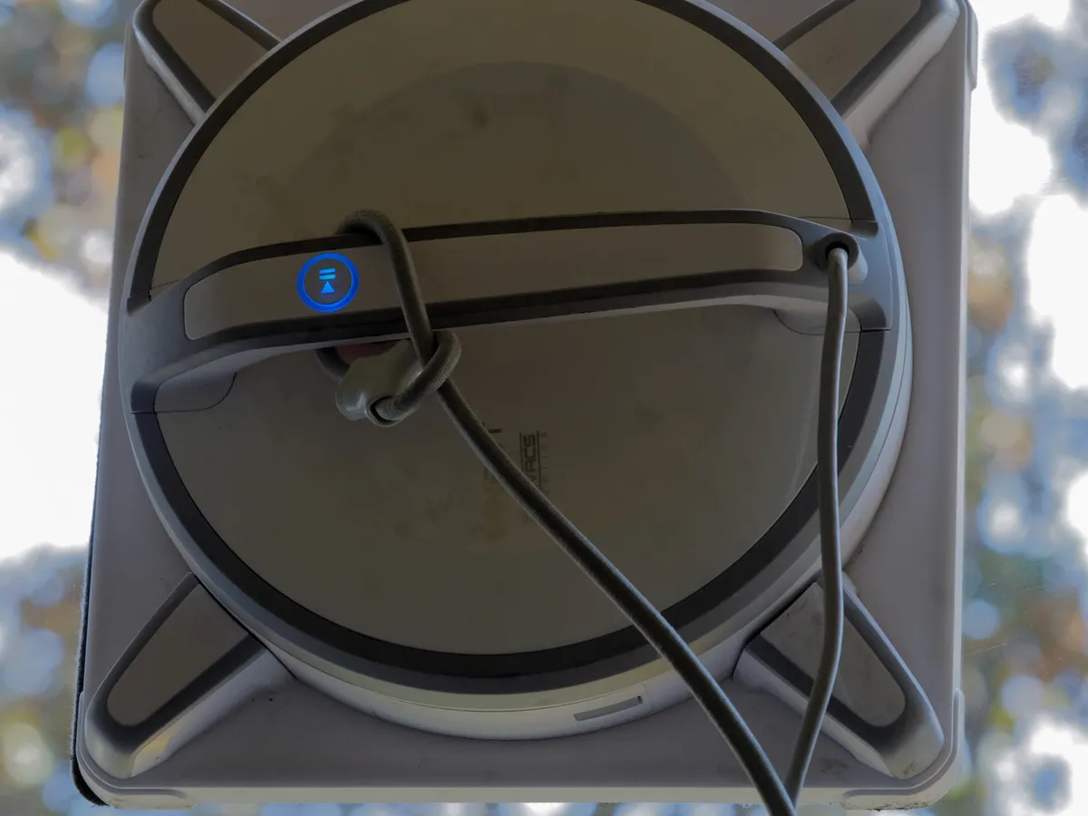An Ecovacs Winbot window-cleaning robot stuck to a window against the light, with its grey safety rope looped through the carrying handle