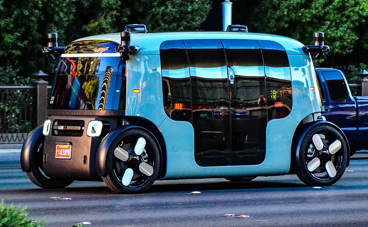 A light teal Zoox robotaxi, a boxy purpose-built electric vehicle with sliding doors, large windows and a sensor pod at each corner of the roof, driving on the Las Vegas Strip